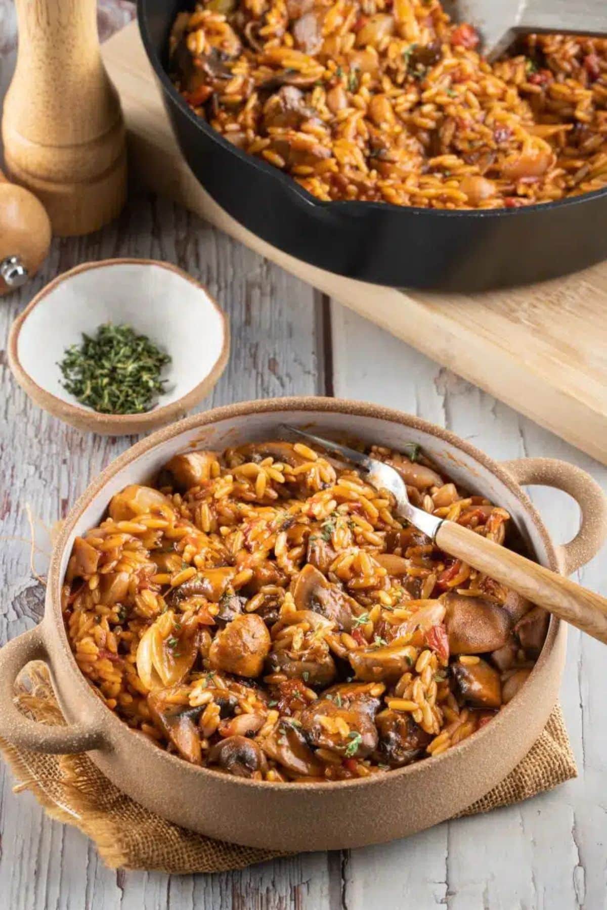 A bowl of orzo pasta with mushrooms and herbs, with a skillet in the background.