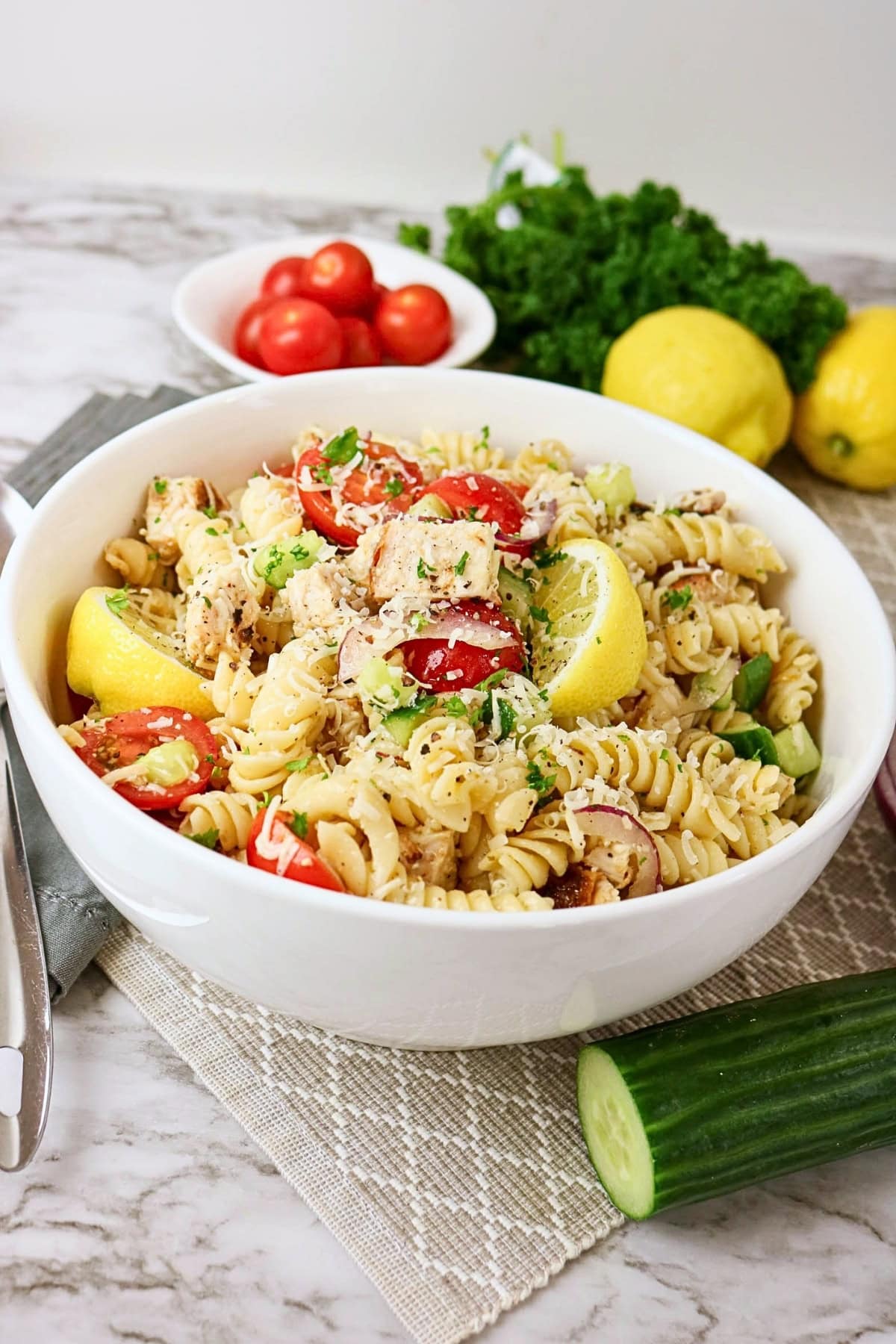 A bowl of rotini pasta salad with chicken, tomatoes, lemon wedges, and herbs.