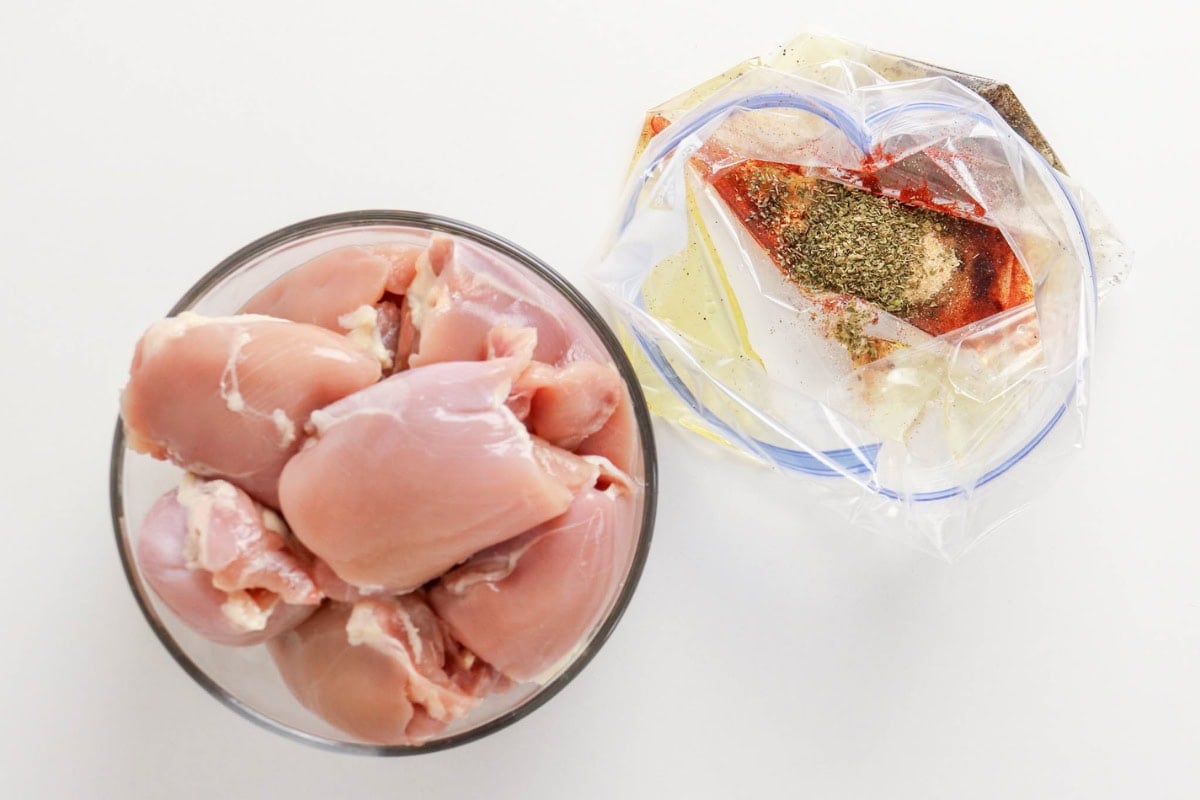 Raw chicken in a bowl next to a zip bag with oil and spices.