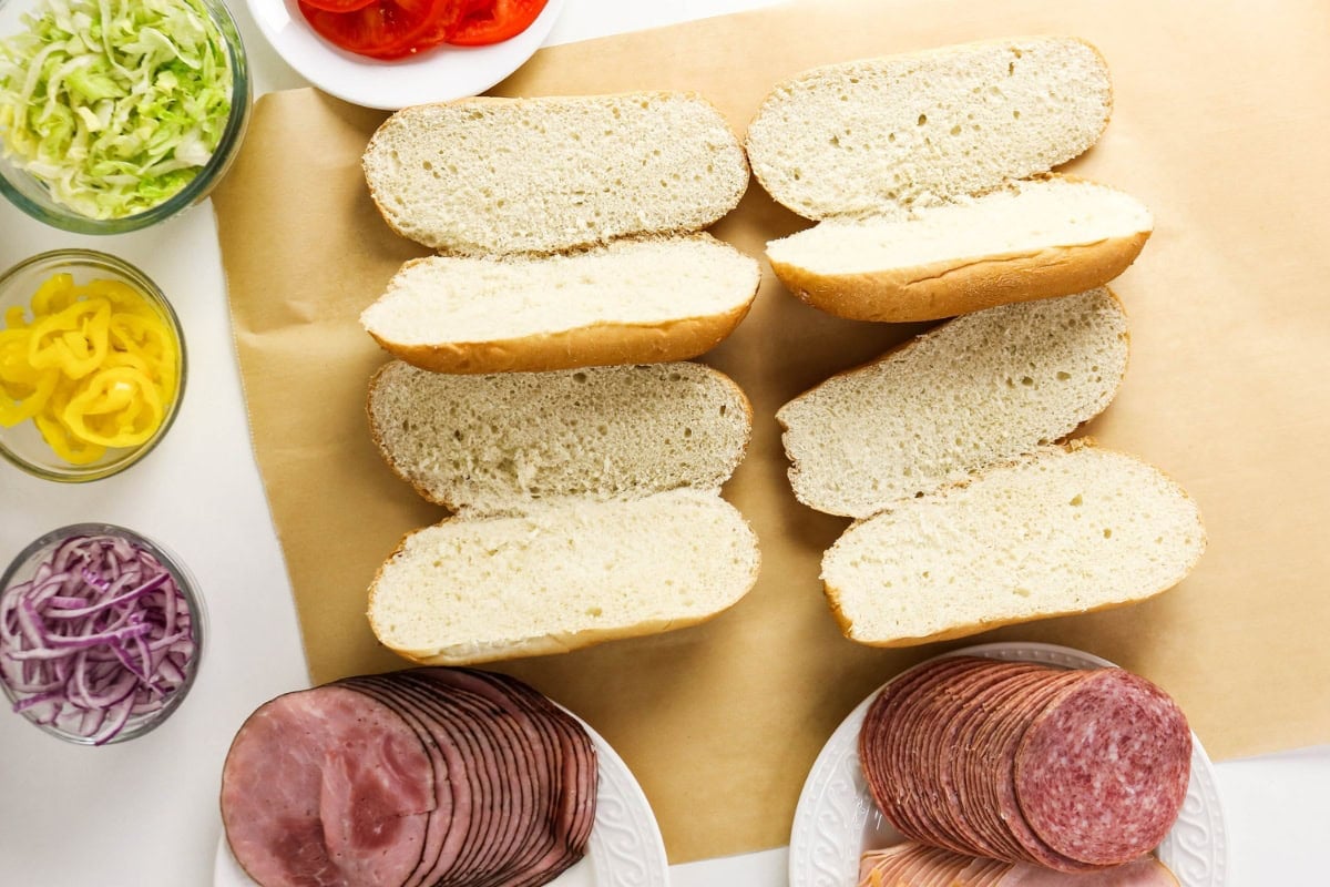 Open hoagie rolls with deli meats, sliced veggies, and toppings arranged around them.