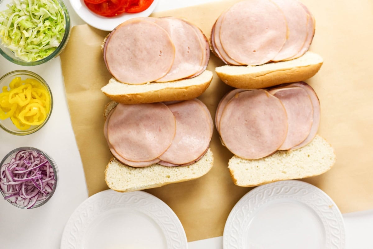 Four hoagie rolls with sliced deli meat, surrounded by bowls of veggies.