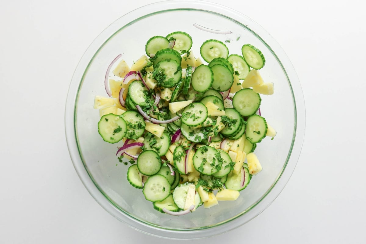 A glass bowl of cucumber, pineapple, red onion, and herbs salad on a white surface.