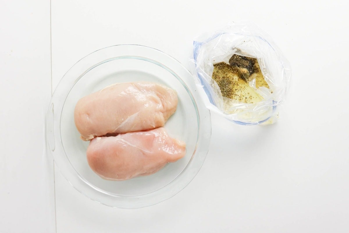 Two raw chicken breasts on a plate next to a bag with marinade and spices.