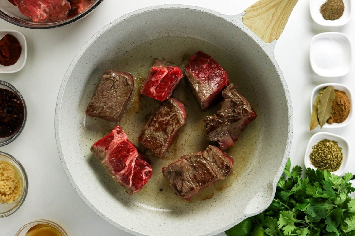 Chunks of beef searing in a white pan, surrounded by bowls of spices and herbs.