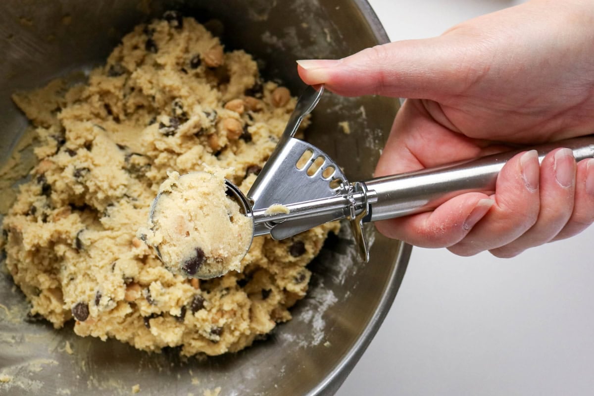 A hand scoops cookie dough with chocolate chips using a metal cookie scoop.
