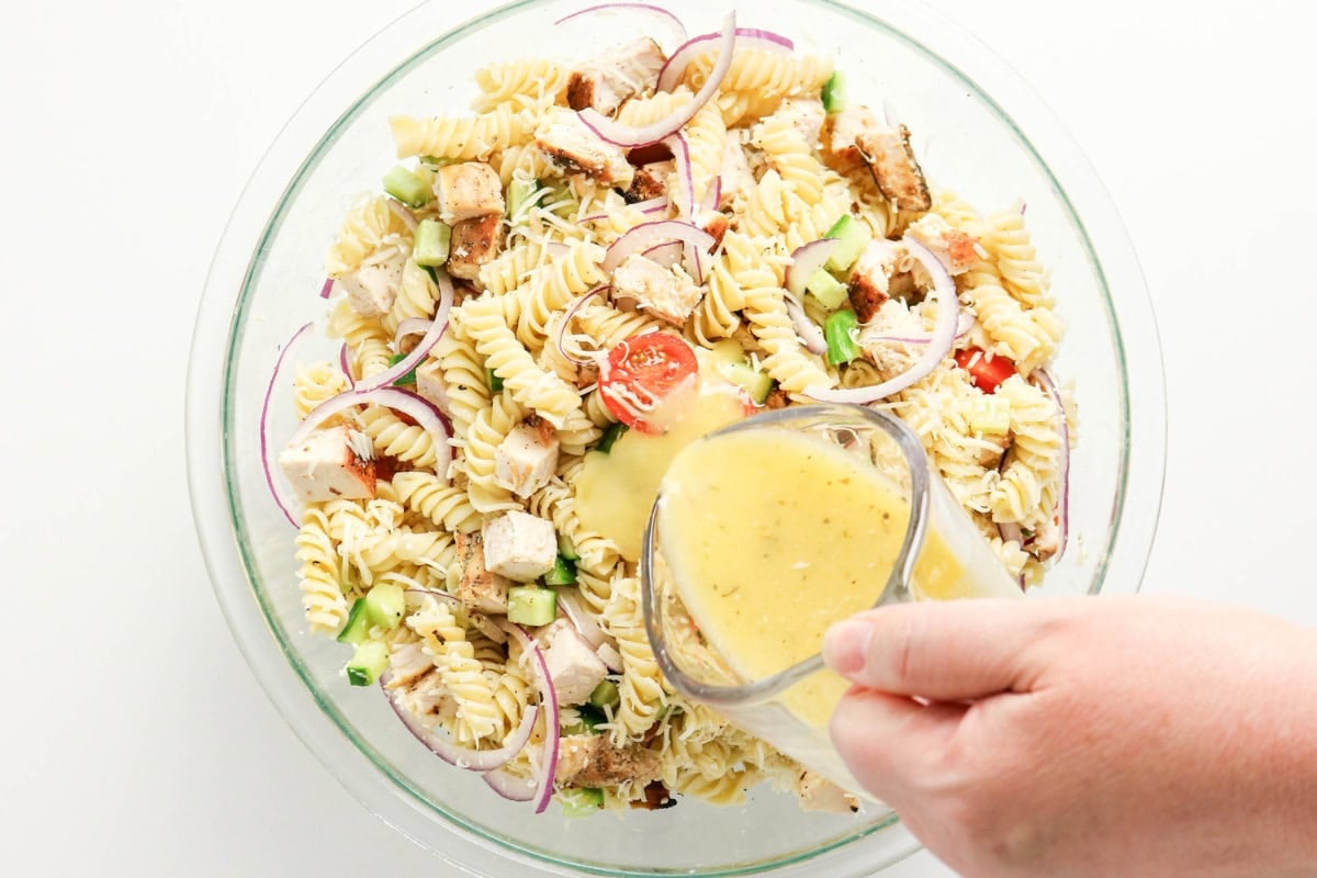 A hand pours dressing onto a bowl of pasta salad with vegetables and chicken.