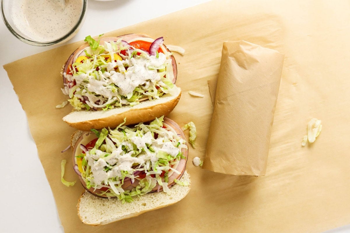 Open sub sandwich with lettuce and dressing beside a wrapped sandwich on parchment.