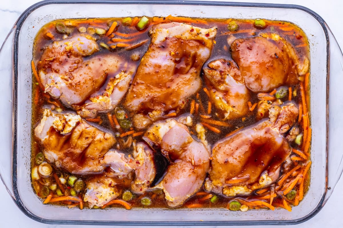 Raw chicken marinating in a glass dish with sauce, carrots, and green onions.