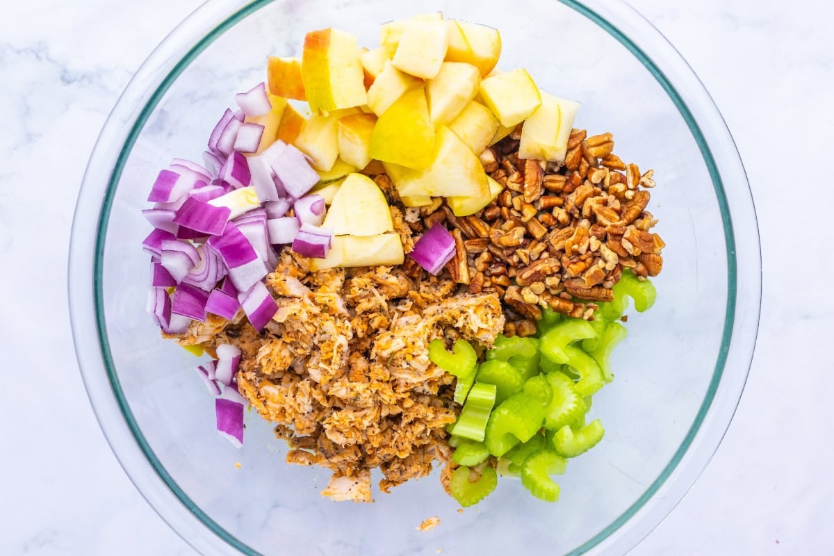Glass bowl with chopped red onion, apple, celery, pecans, and shredded chicken.