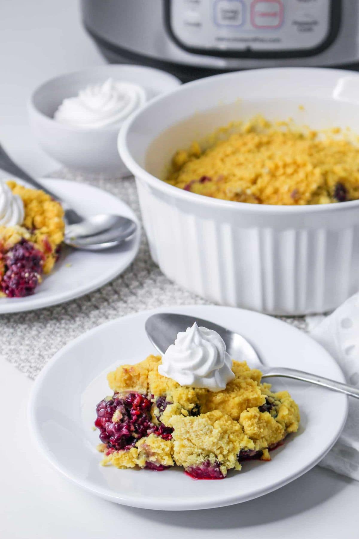 Mixed berry cobbler topped with whipped cream on white plates, with a spoon.