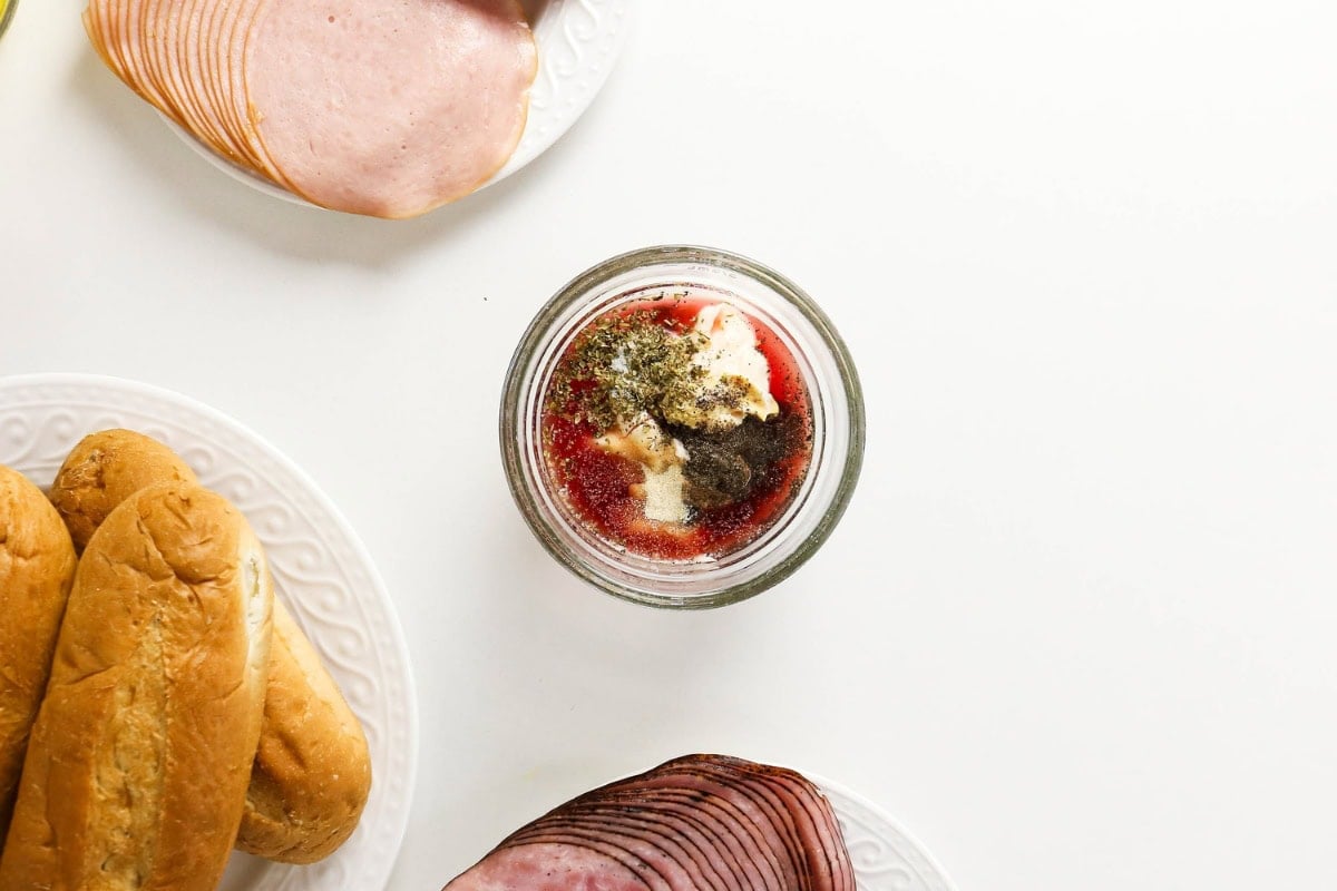Jar of sandwich spread, deli meats, and buns on a white surface.