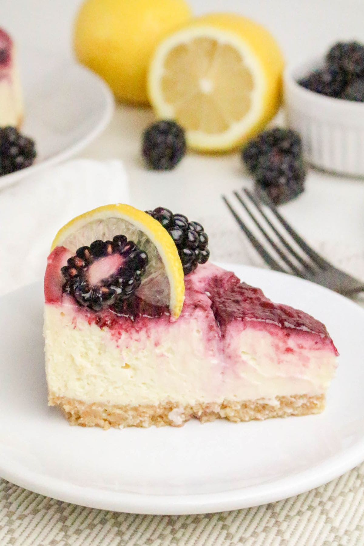 A slice of cheesecake topped with blackberries and a lemon slice on a white plate.
