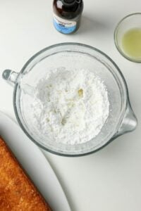 Glass bowl with powdered sugar, lemon juice, vanilla, and part of a baked cake.