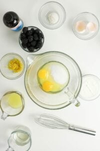 Baking ingredients including eggs, sugar, lemon, blackberries, and a whisk on a table.