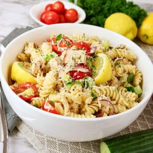 A bowl of pasta salad with chicken, tomatoes, lemon wedges, and herbs.