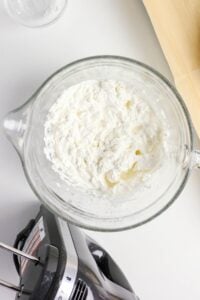 A bowl of whipped cream next to a hand mixer on a white surface.