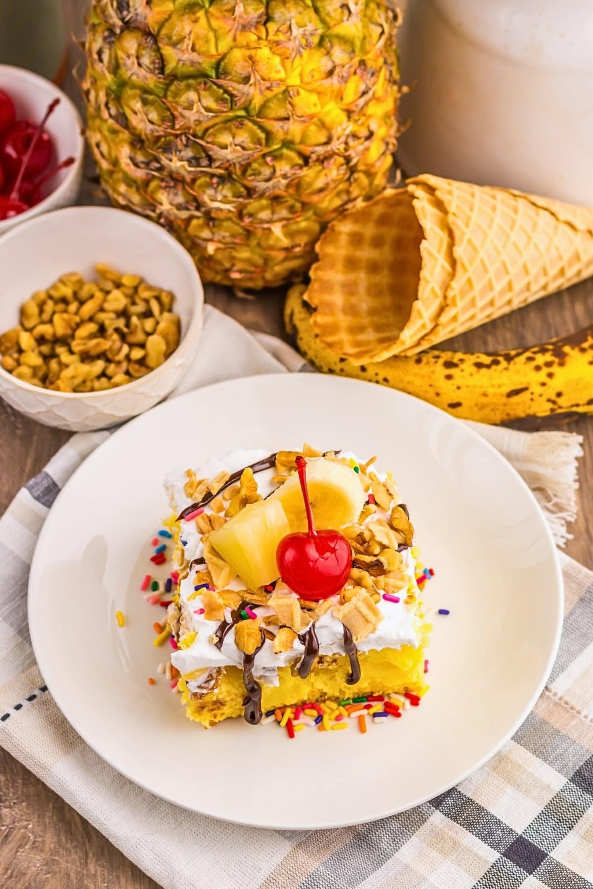 A slice of banana split cake topped with pineapple, cherry, nuts, and sprinkles.