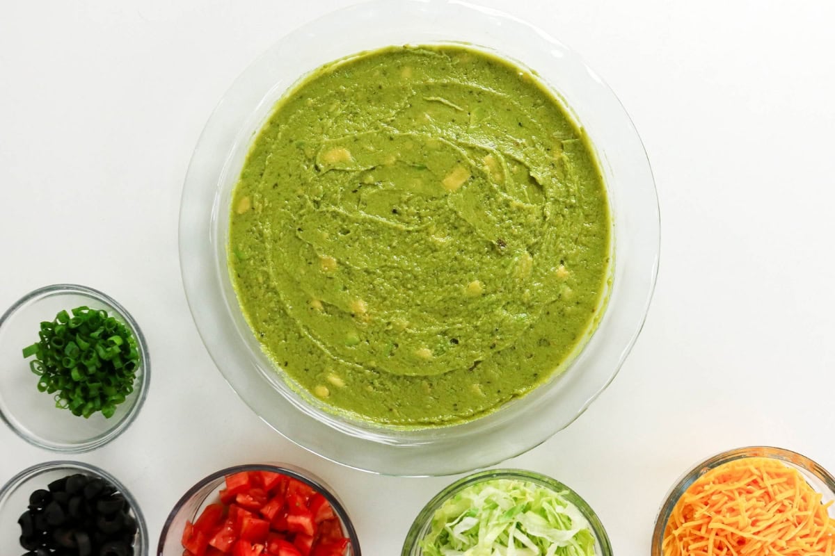 A bowl of guacamole surrounded by bowls of toppings on a white background.