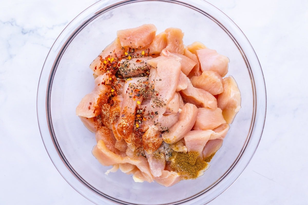Raw chicken pieces in a glass bowl with spices sprinkled on top.