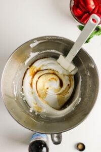 Metal mixing bowl with whipped cream, vanilla, and a spatula on a white surface.