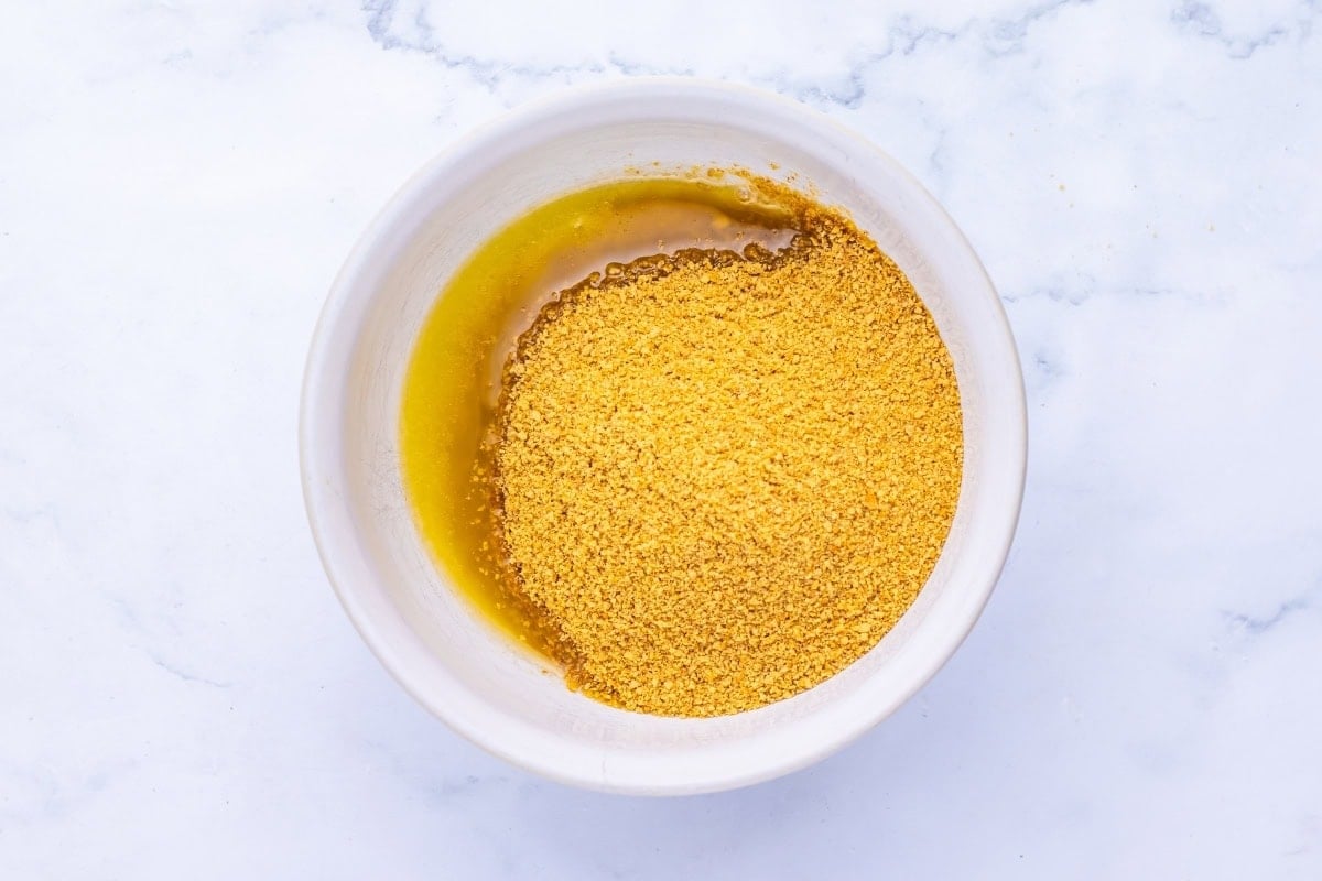 A white bowl with crushed graham crackers and melted butter on a marble surface.