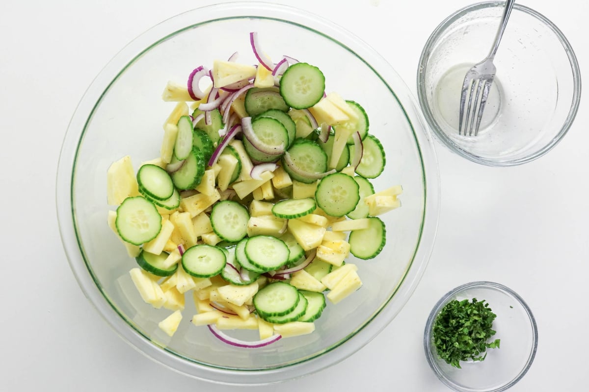 A glass bowl with sliced cucumber, pineapple, and red onion on a white surface.