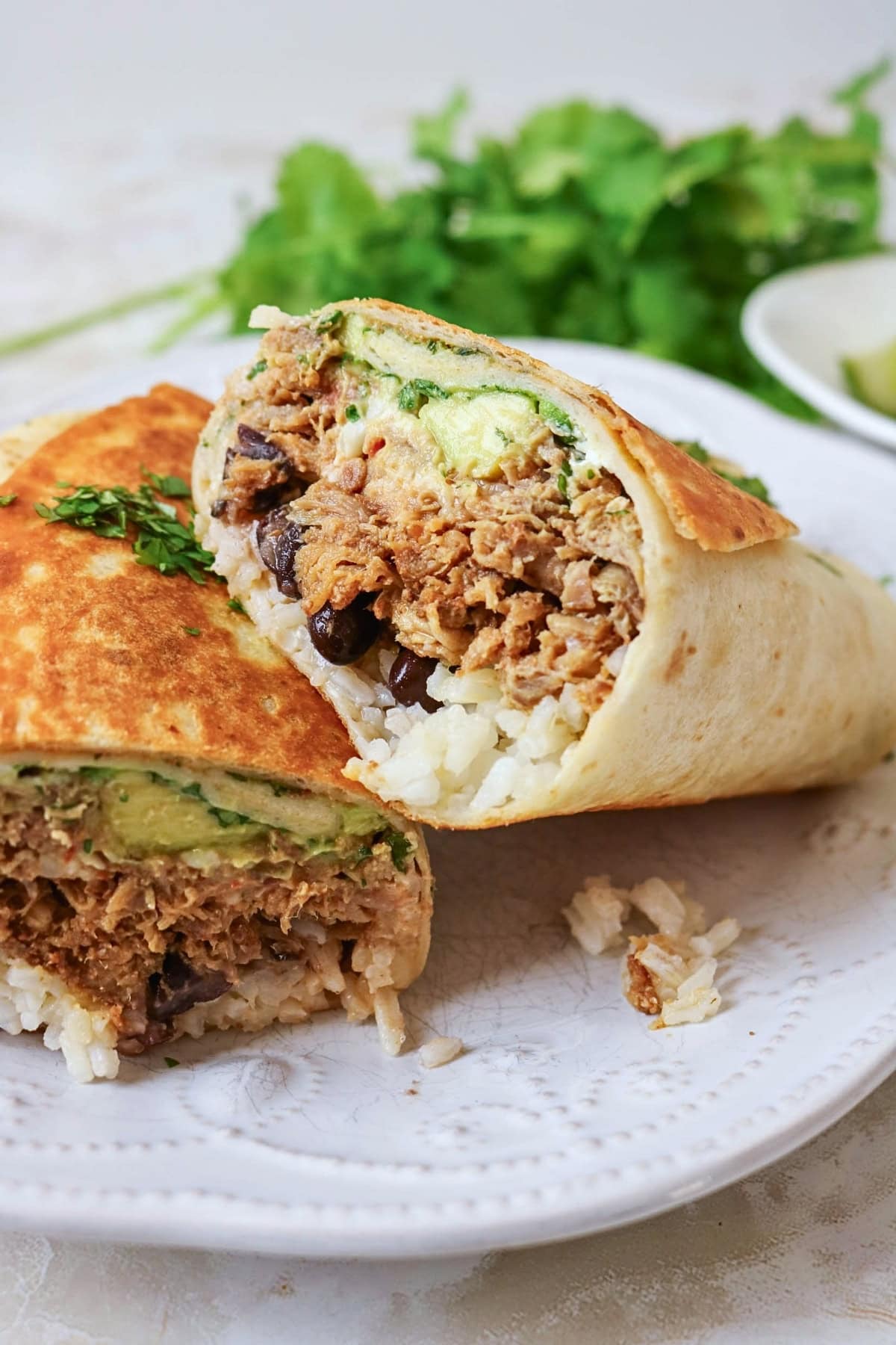 A burrito cut in half, filled with rice, beans, meat, and avocado on a plate.