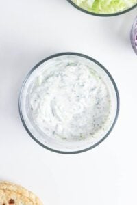 A bowl of creamy white tzatziki sauce with herbs on a white surface.