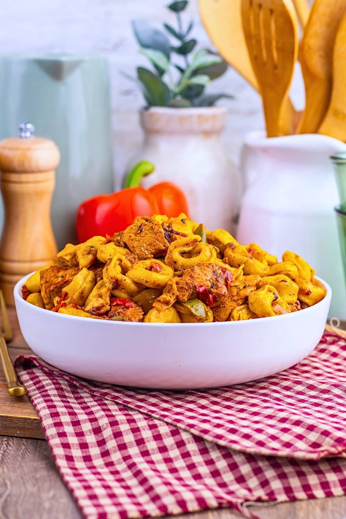 A bowl of pasta with vegetables on a red checkered cloth, kitchen items in background.