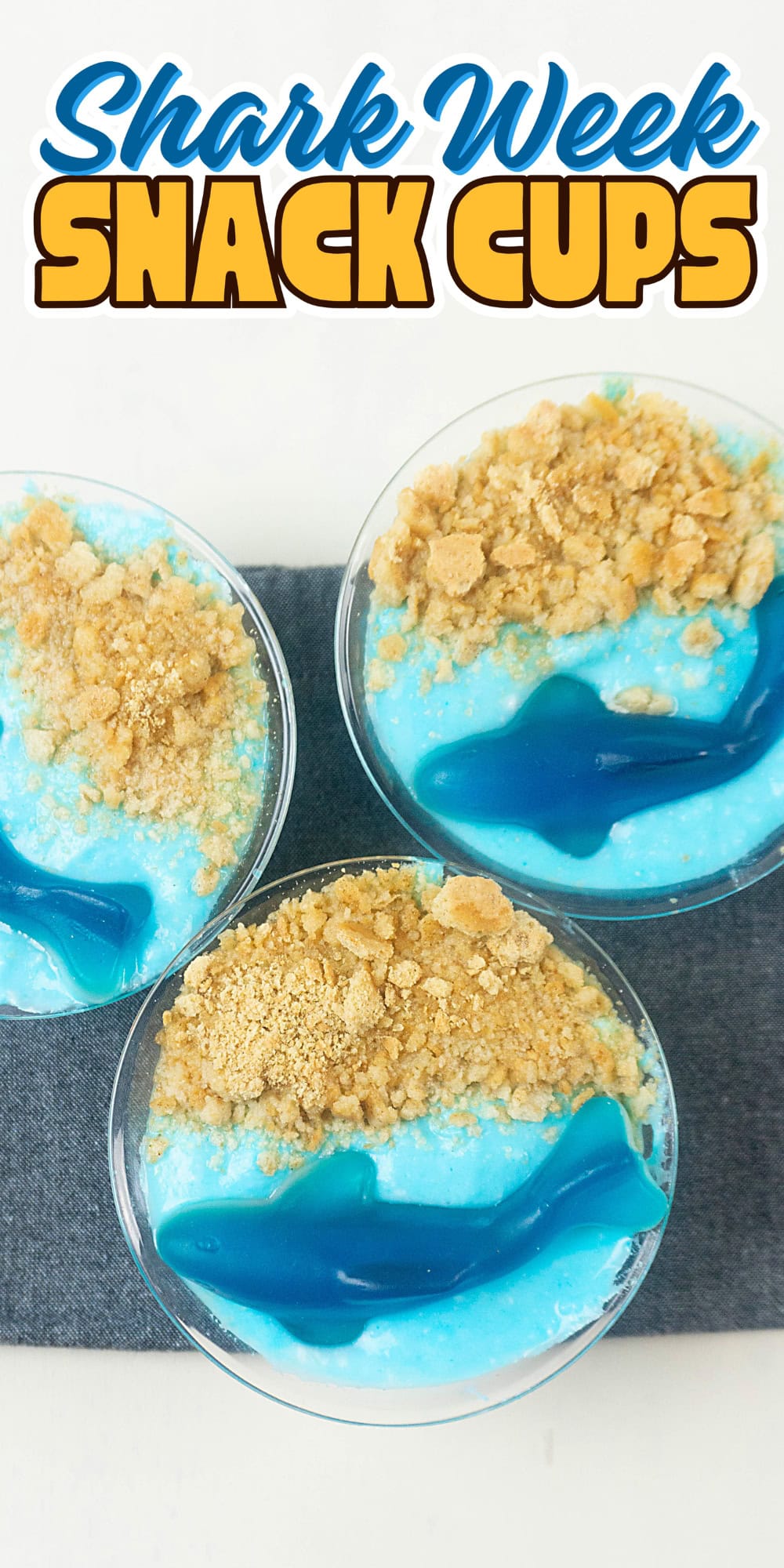 Three blue snack cups topped with cookie crumbs and gummy sharks on a white surface.