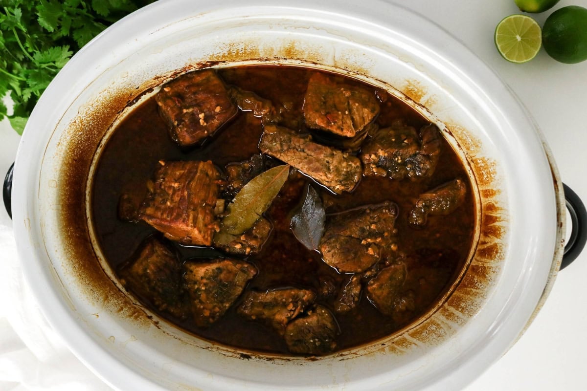 Chunks of beef simmering in a rich, dark sauce in a white slow cooker.