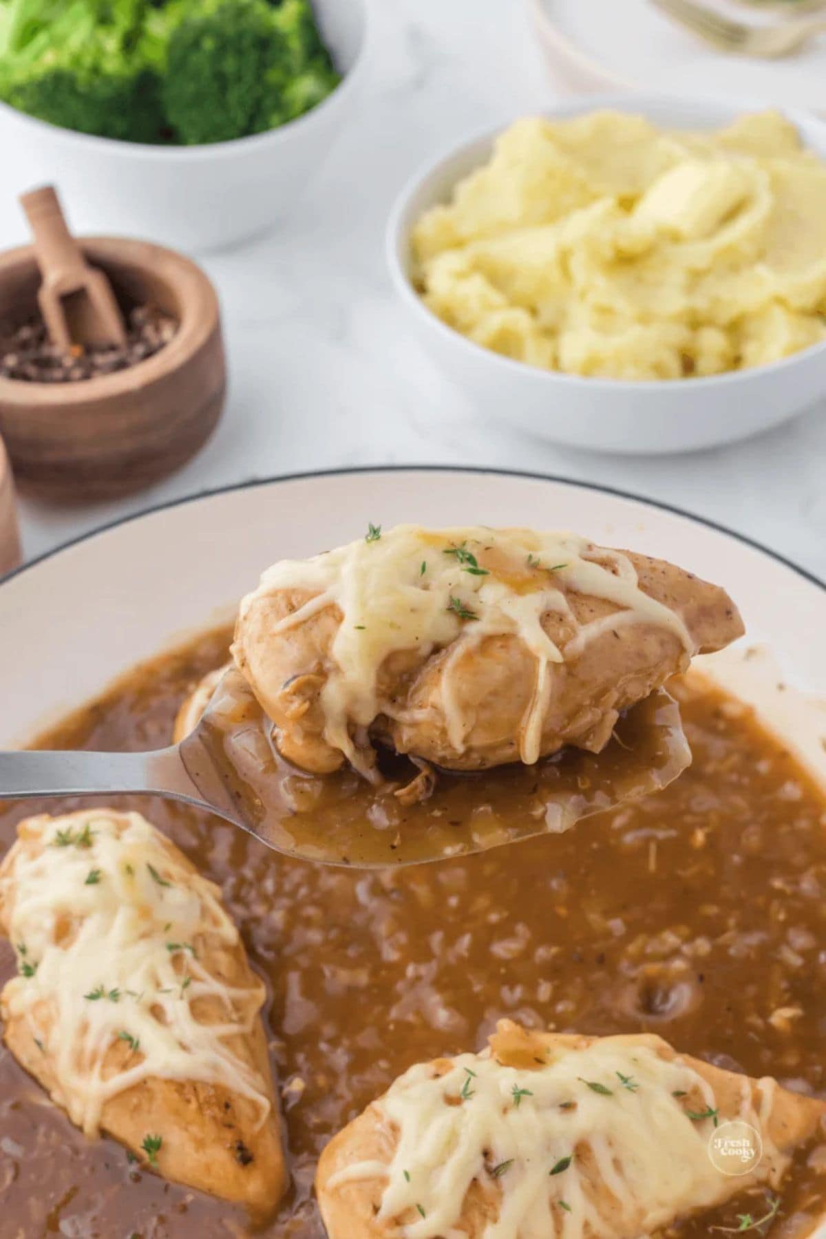 Chicken breast with melted cheese in brown gravy, with mashed potatoes and broccoli.