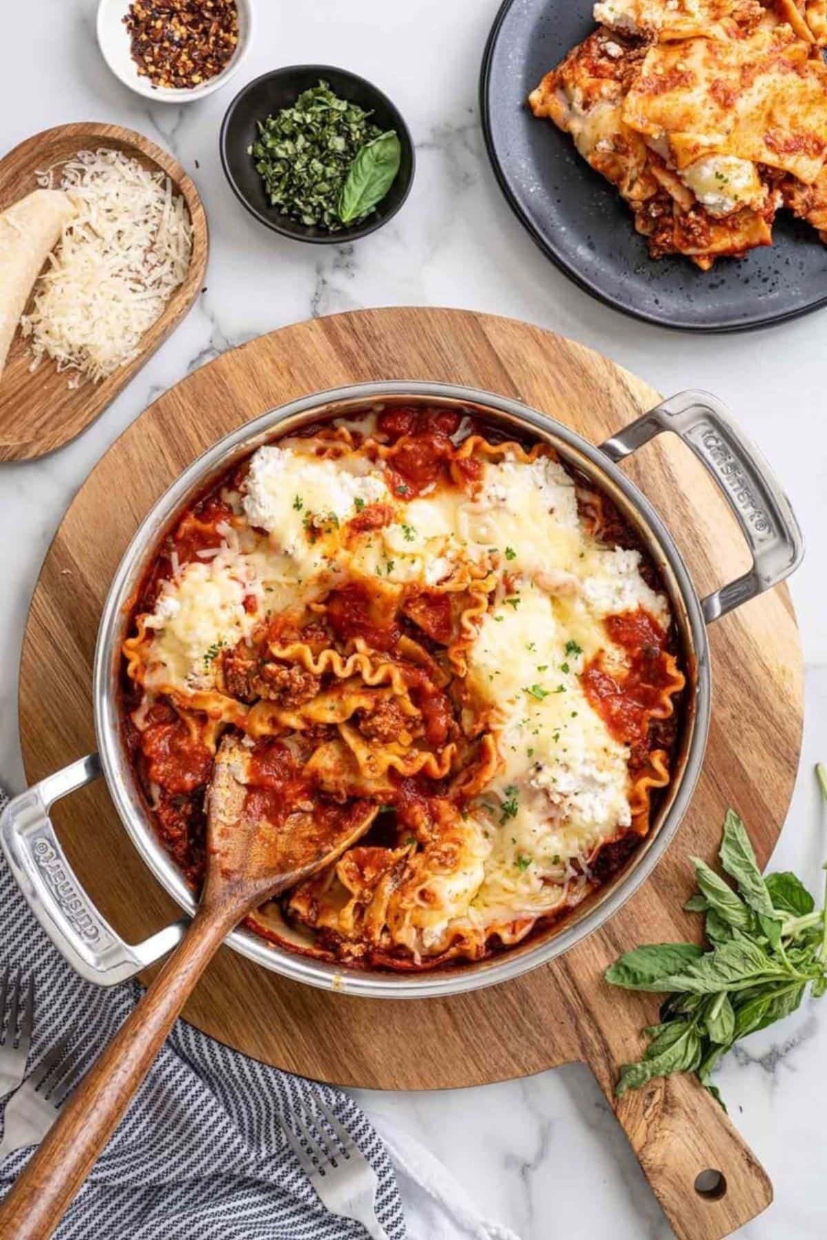 Pan of cheesy lasagna with tomato sauce, herbs, and a wooden spoon on the side.