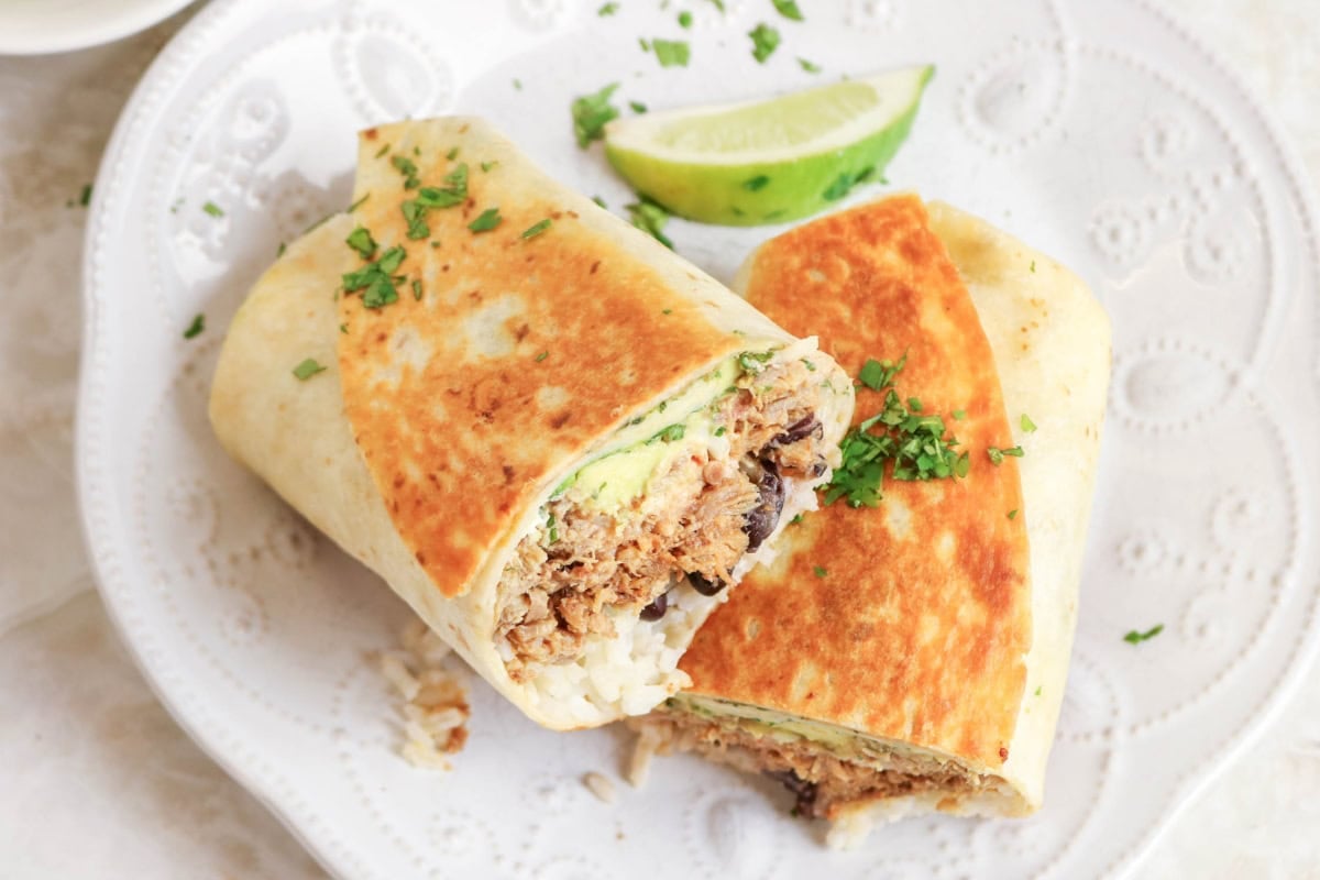 A burrito cut in half on a plate with a lime wedge and chopped herbs on top.