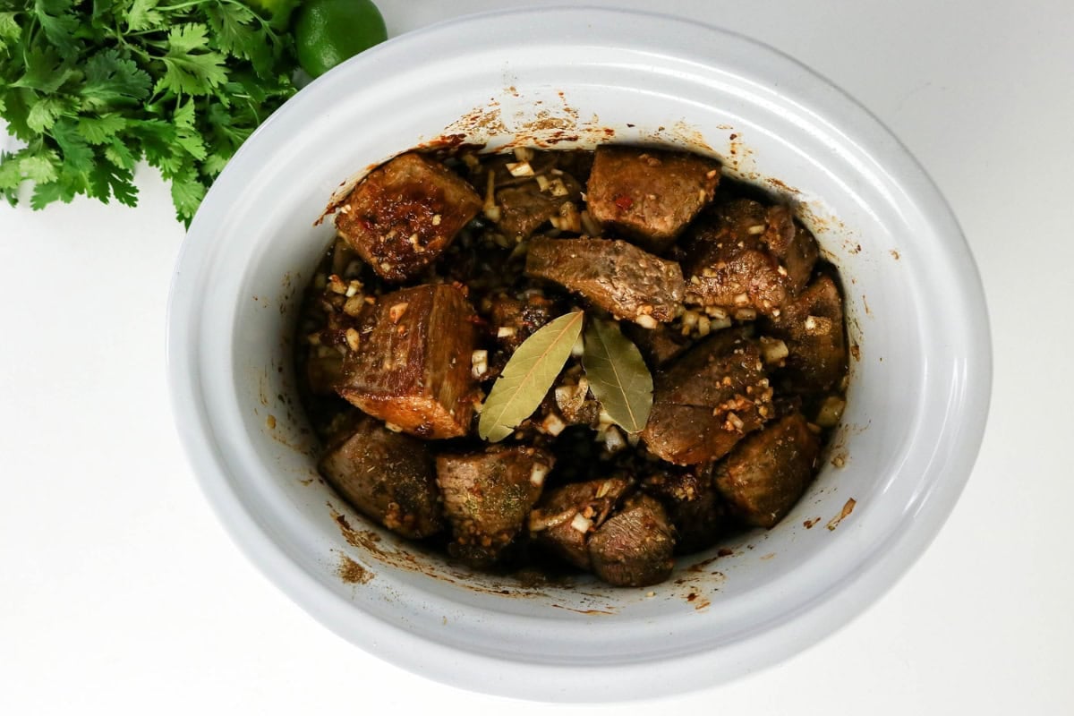 Chunks of seasoned beef and bay leaves in a slow cooker, with herbs on the side.