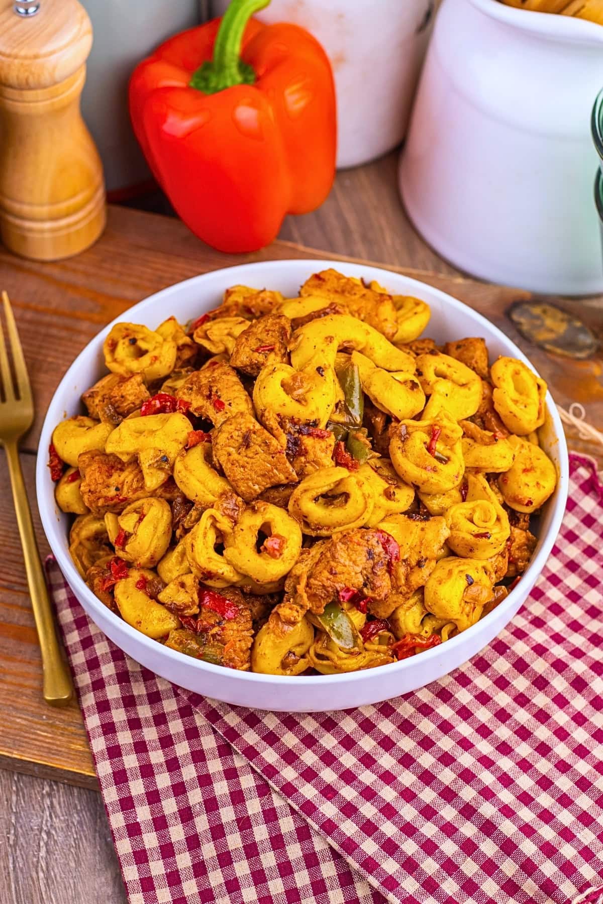 A bowl of tortellini pasta with chicken and peppers on a red checkered cloth.