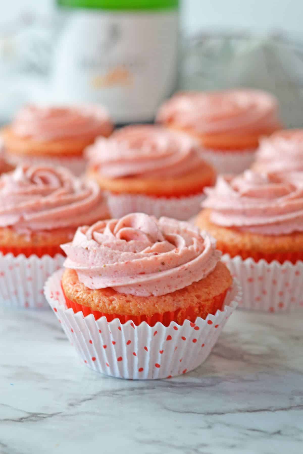 Pink cupcakes with swirled frosting in white polka dot wrappers on a marble surface.