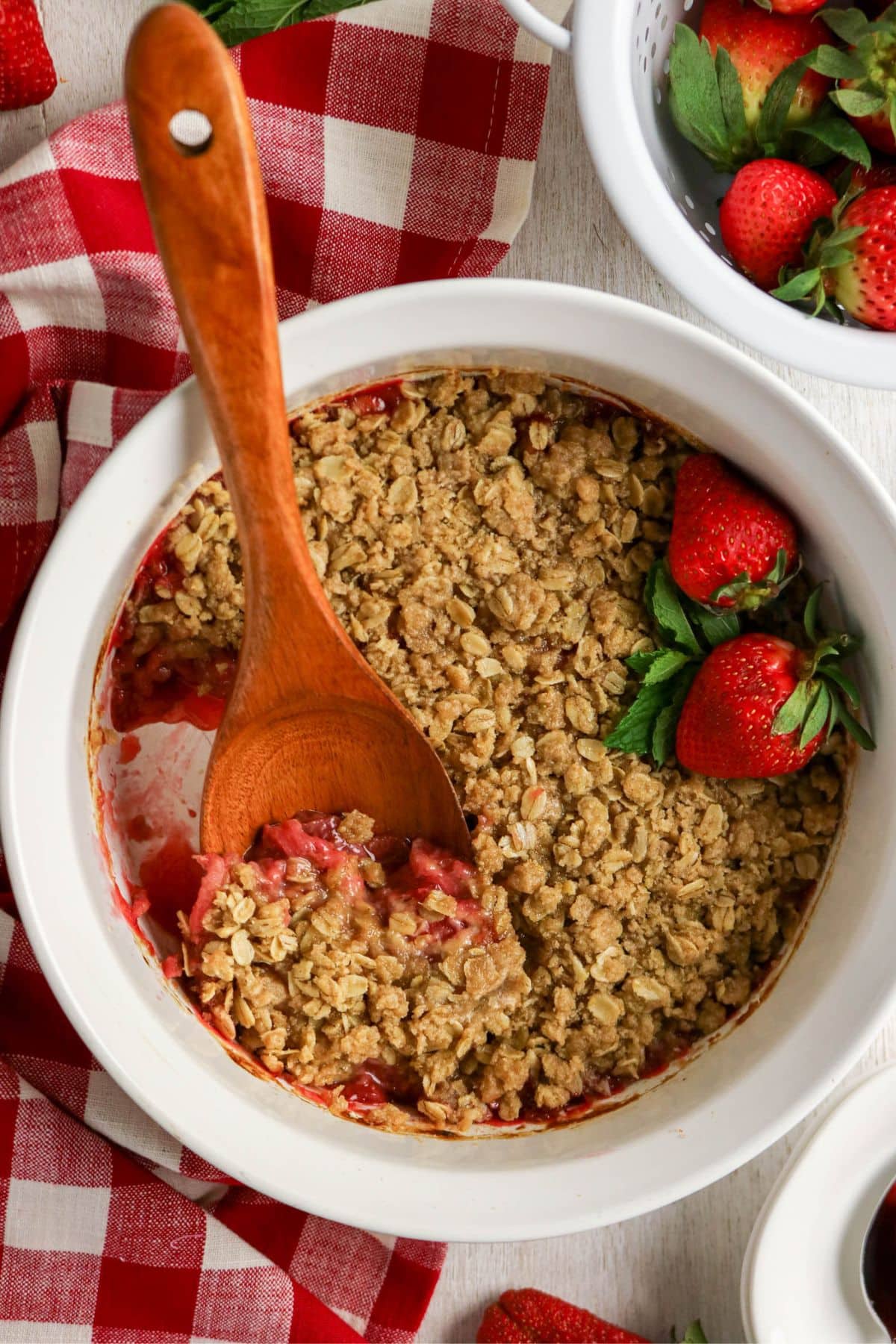 Baked strawberry crisp in a white dish with a wooden spoon and fresh strawberries.