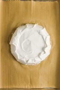 Round cake topped with swirled white frosting on parchment paper.