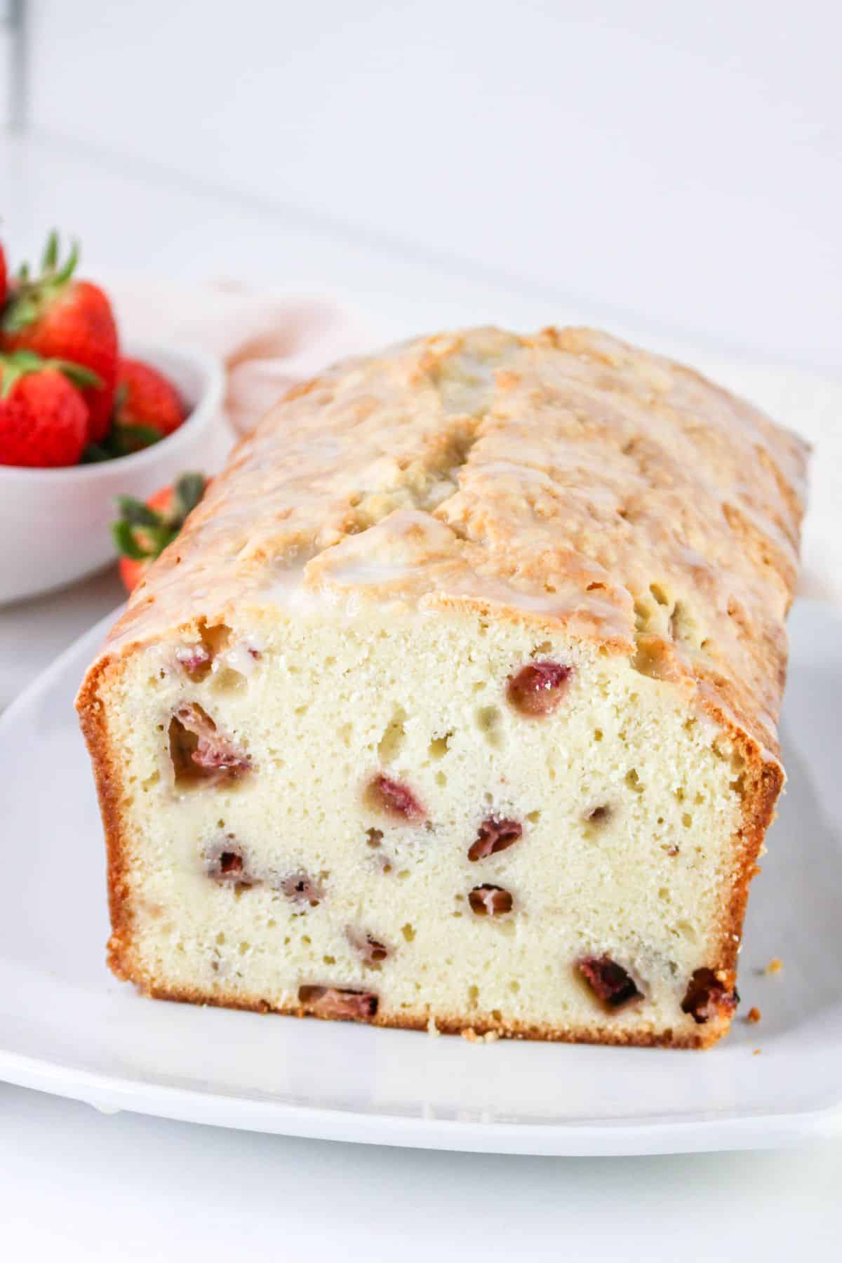 A sliced loaf of bread with fruit pieces, on a white plate near strawberries.