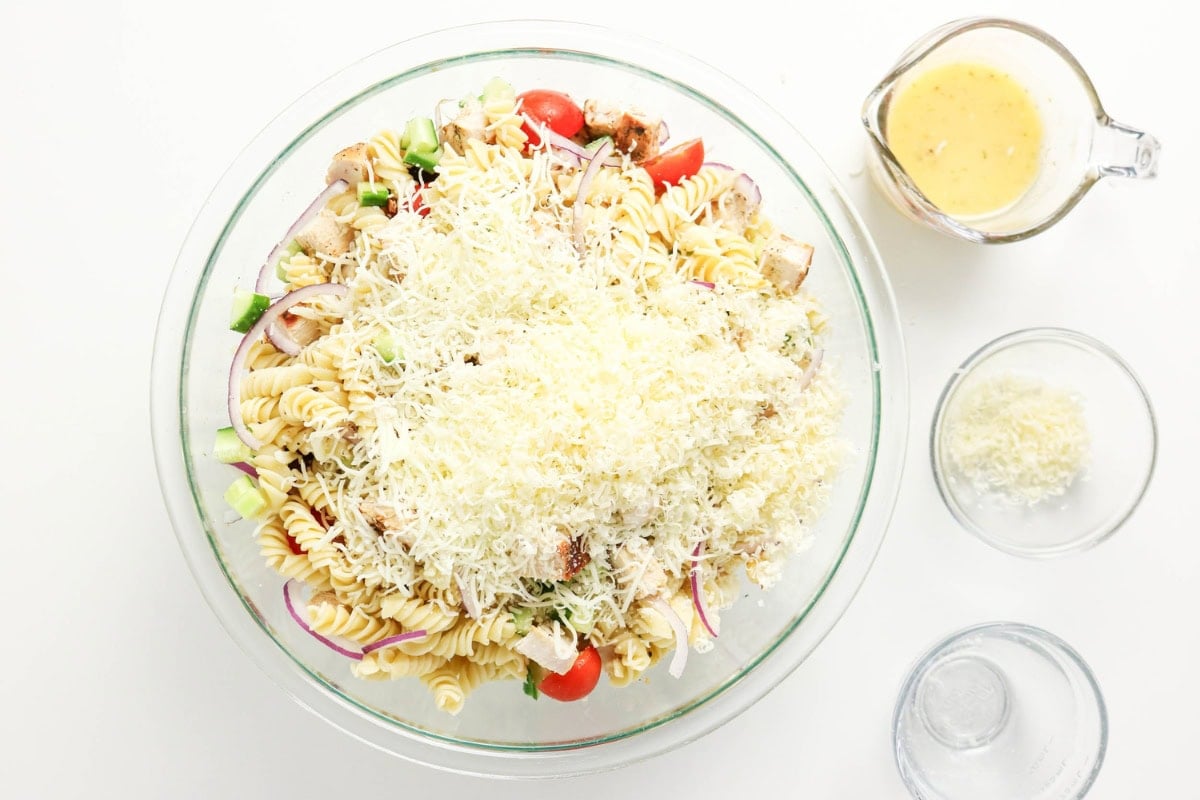 A glass bowl of pasta salad topped with shredded cheese, beside dressing and cheese.