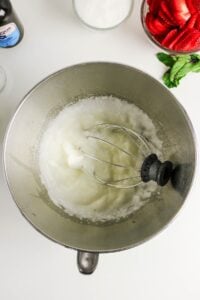 Metal mixing bowl with whisk, whipped egg whites, strawberries, and sugar nearby.