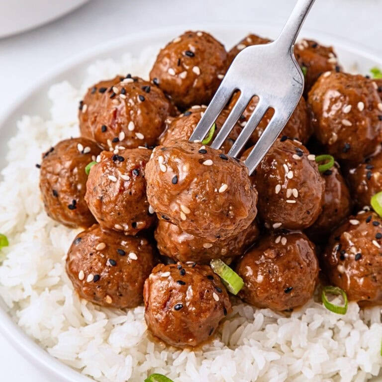 Fork picking up saucy meatballs with sesame seeds over white rice on a plate.