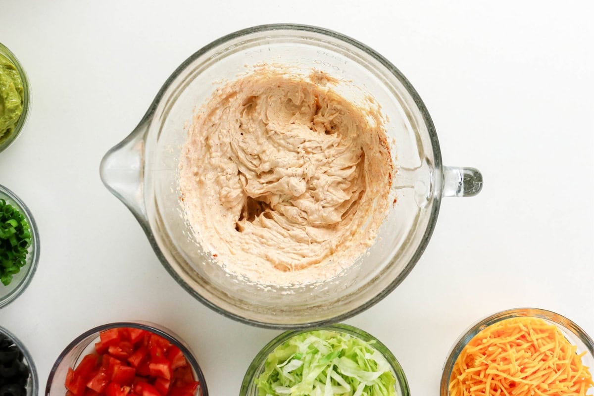 A glass bowl of creamy dip surrounded by bowls of chopped taco toppings.