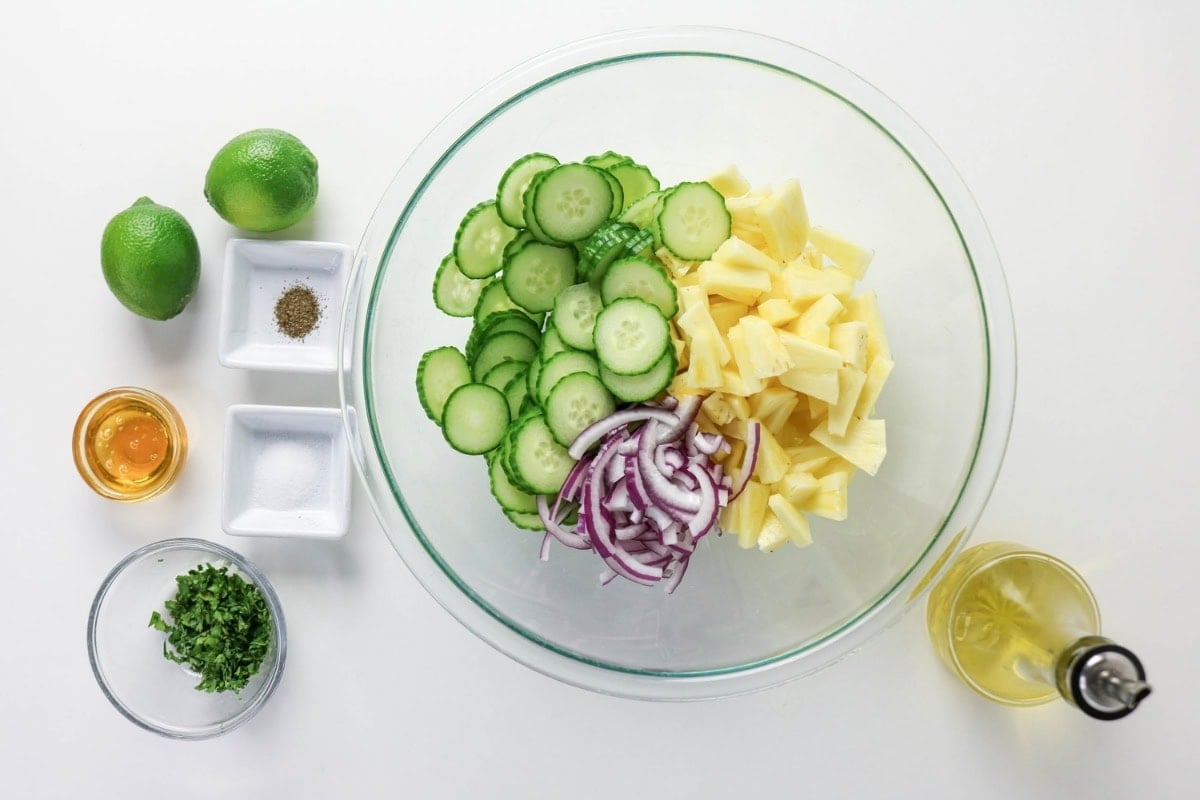 Sliced cucumber, pineapple, and onion in a bowl with salad ingredients nearby.