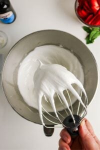 A whisk holds stiff glossy whipped meringue above a mixing bowl.
