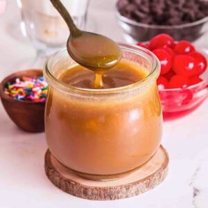 A jar of caramel sauce with a spoon, cherries, sprinkles, and chocolate behind.