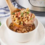 A wooden spoon serves black-eyed peas with ham into a white bowl.