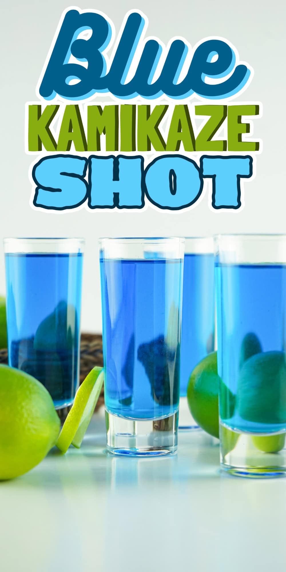 Four blue kamikaze shots surrounded by fresh limes on a white surface.
