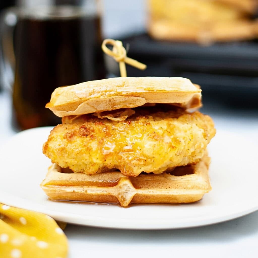 A chicken waffle sandwich on a white plate with a skewer on top.